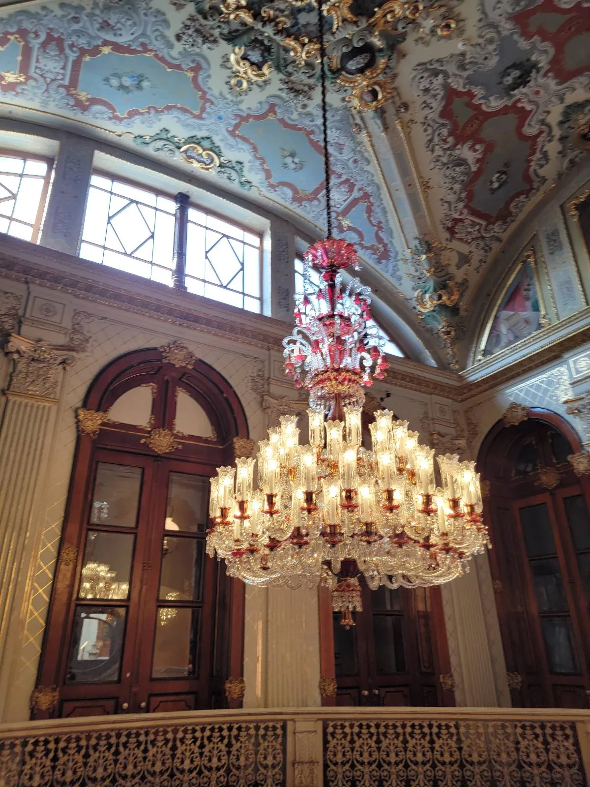 An ornate chandelier with numerous lights hangs from a decorative ceiling in an elegant room with arched windows.