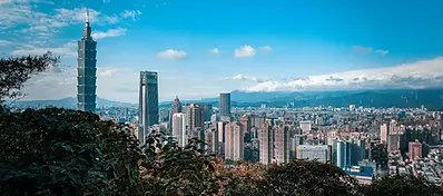 A panoramic view of the Taipei skyline from a high vantage point, dominated by the Taipei 101 skyscraper under a clear blue sky with mountains in the distance.
