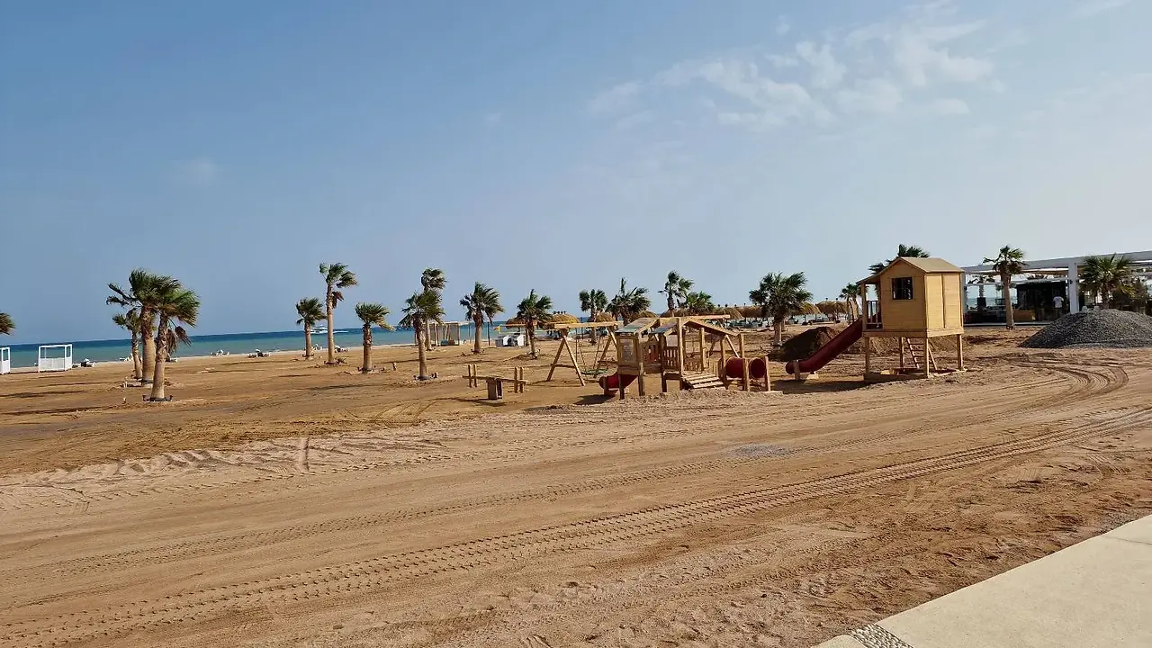A sandy beach with climbing structures and slides is set near a beach with palm trees and the ocean in the background under a partly cloudy sky.