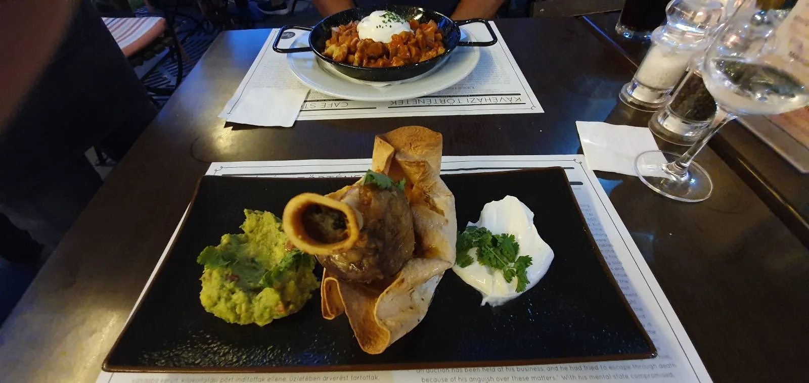 A black rectangular plate holds a dish with a crispy, rolled tortilla filled with a savory filling, accompanied by guacamole and sour cream on the side. In the background, another dish is visible on a white plate. A glass of wine is on the table.