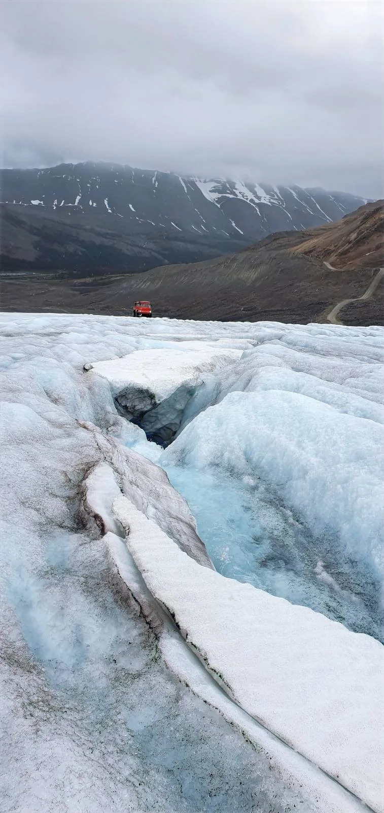 A deep crevasse cuts through a glacier in the foreground, with a small red figure in the distance. Snowy mountains and cloudy skies form a dramatic backdrop.