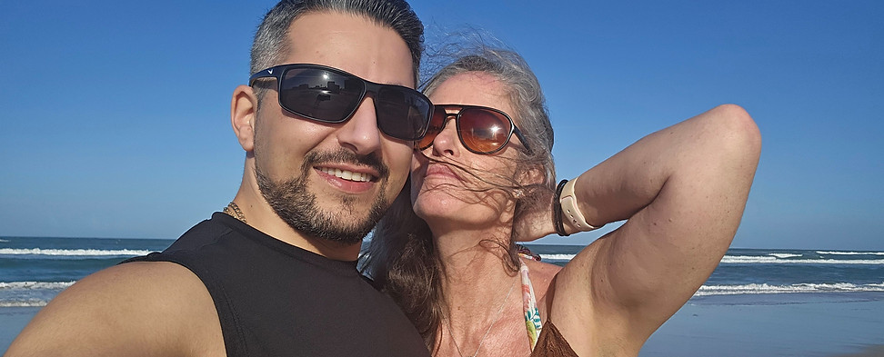 A man and woman wearing sunglasses smile and pose for a selfie on a sunny beach, with the ocean and blue sky in the background. The woman’s hair is blowing in the wind as she holds one hand behind her head.