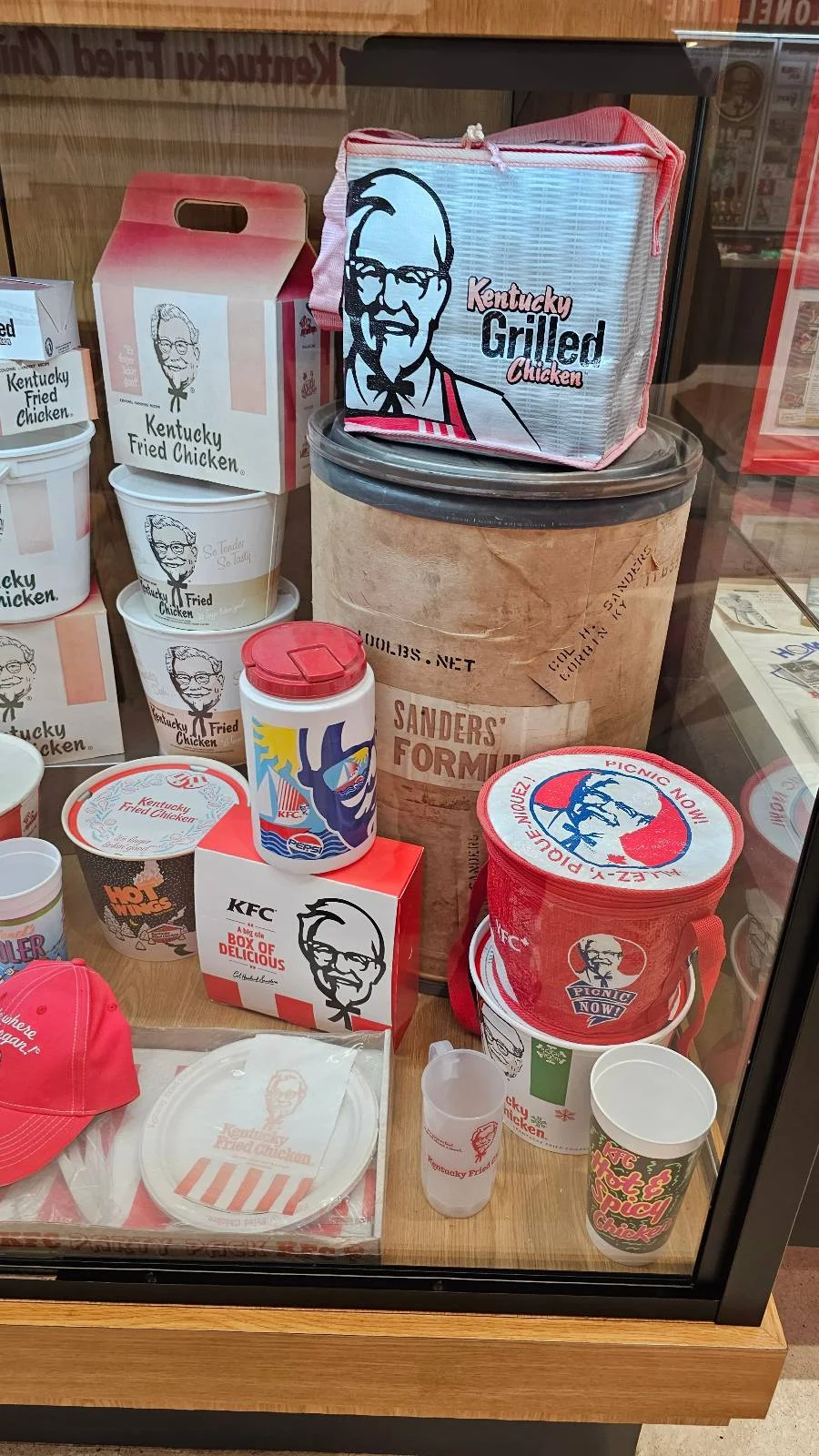 A display case filled with various vintage KFC buckets, boxes, and containers featuring the classic Colonel Sanders logo and red-and-white designs.