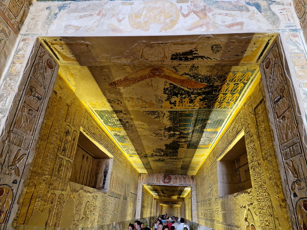 A narrow, ancient Egyptian corridor with a decorated ceiling and walls covered in hieroglyphics. Tourists can be seen walking through the passage.
