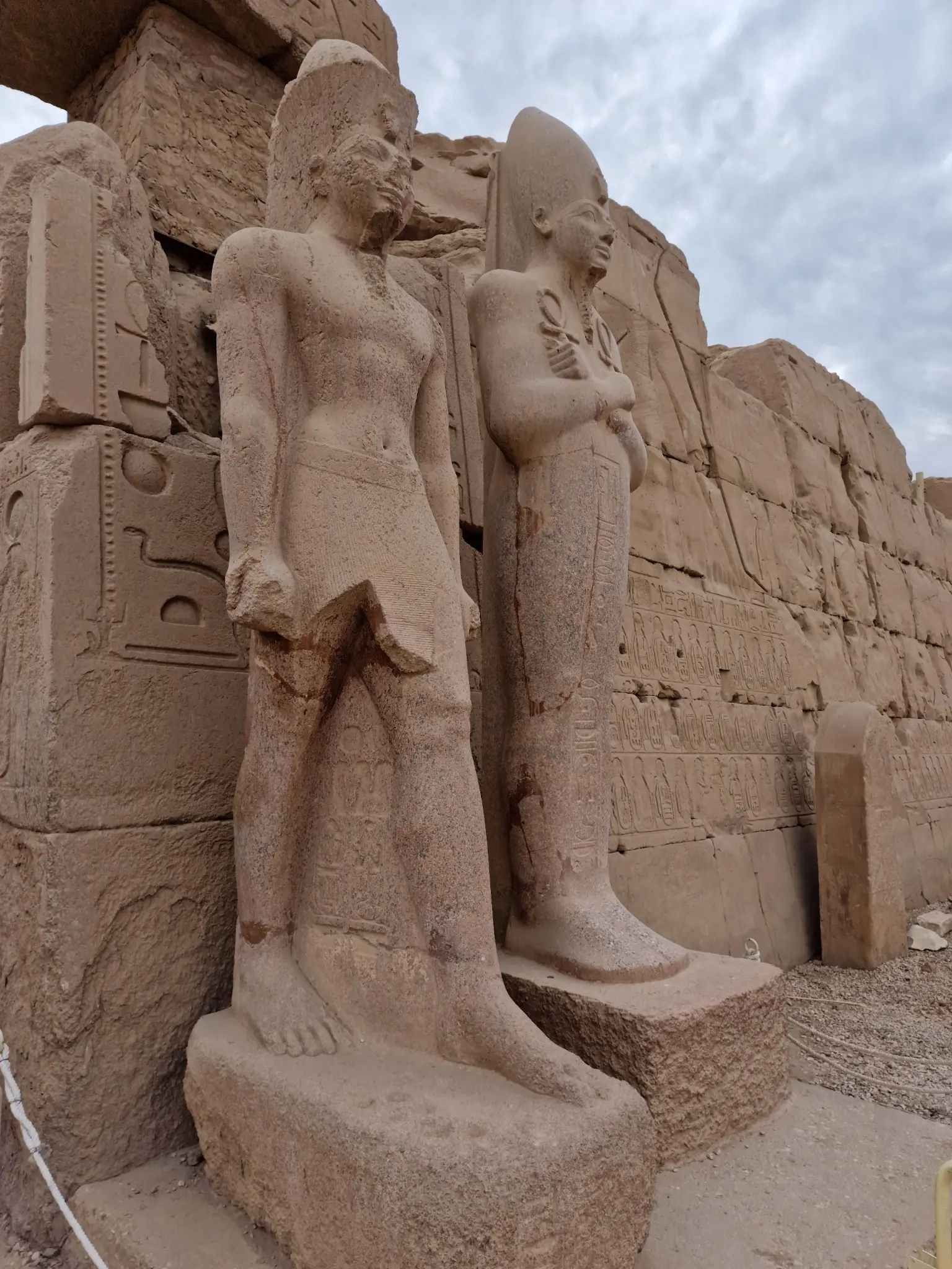 Two large stone statues of ancient Egyptian figures stand side by side in front of a weathered stone wall featuring carved hieroglyphs. The sky above is cloudy.