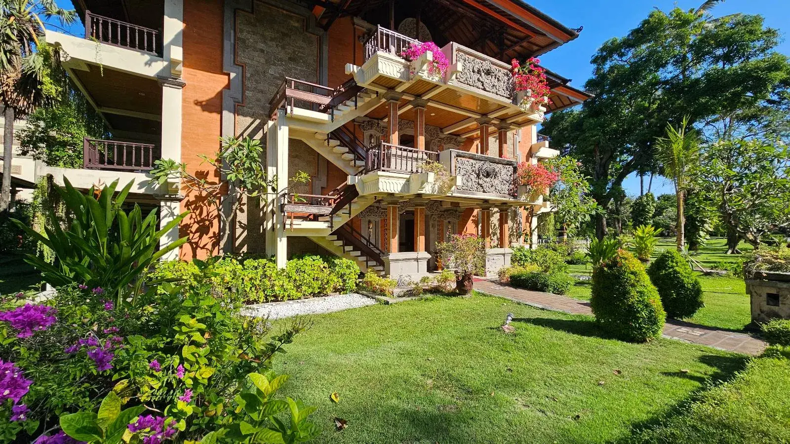 A three-story building with ornate balconies covered in lush greenery and vibrant pink flowers. The structure is surrounded by a well-kept garden with shrubs, grass, and trees under a clear blue sky. A meandering path leads to the entrance.