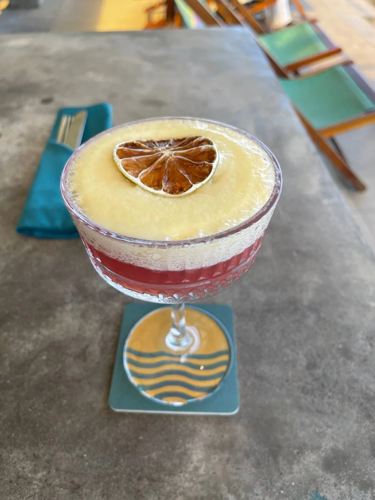 A cocktail in a glass with a frothy cream layer and a dried citrus slice on top. The drink rests on a coaster with a wave design, set on a stone-like surface. A folded blue napkin is in the background.