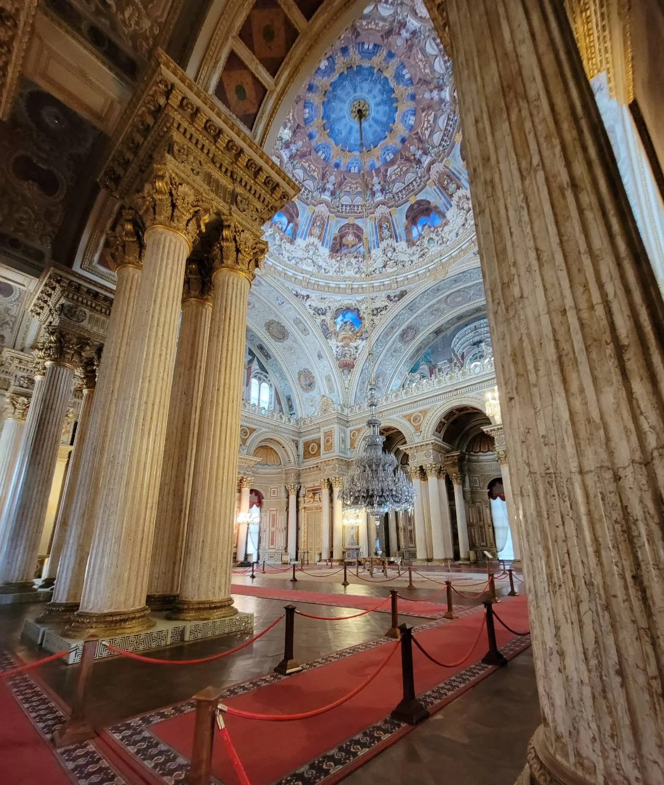 Grand interior of a historic building with tall ornate columns, intricate ceiling artwork, a large patterned dome, red carpet, and rope barriers. Light streams in, highlighting architectural details.