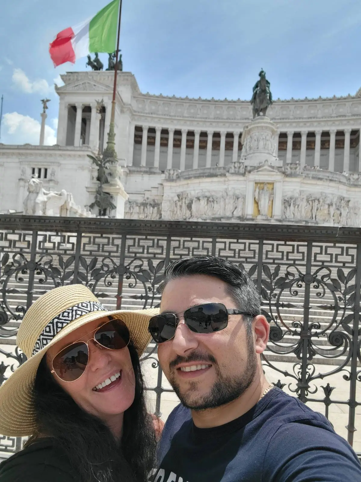A smiling couple wearing sunglasses poses for a selfie in front of a grand white stone building with columns and statues, featuring an Italian flag flying above.