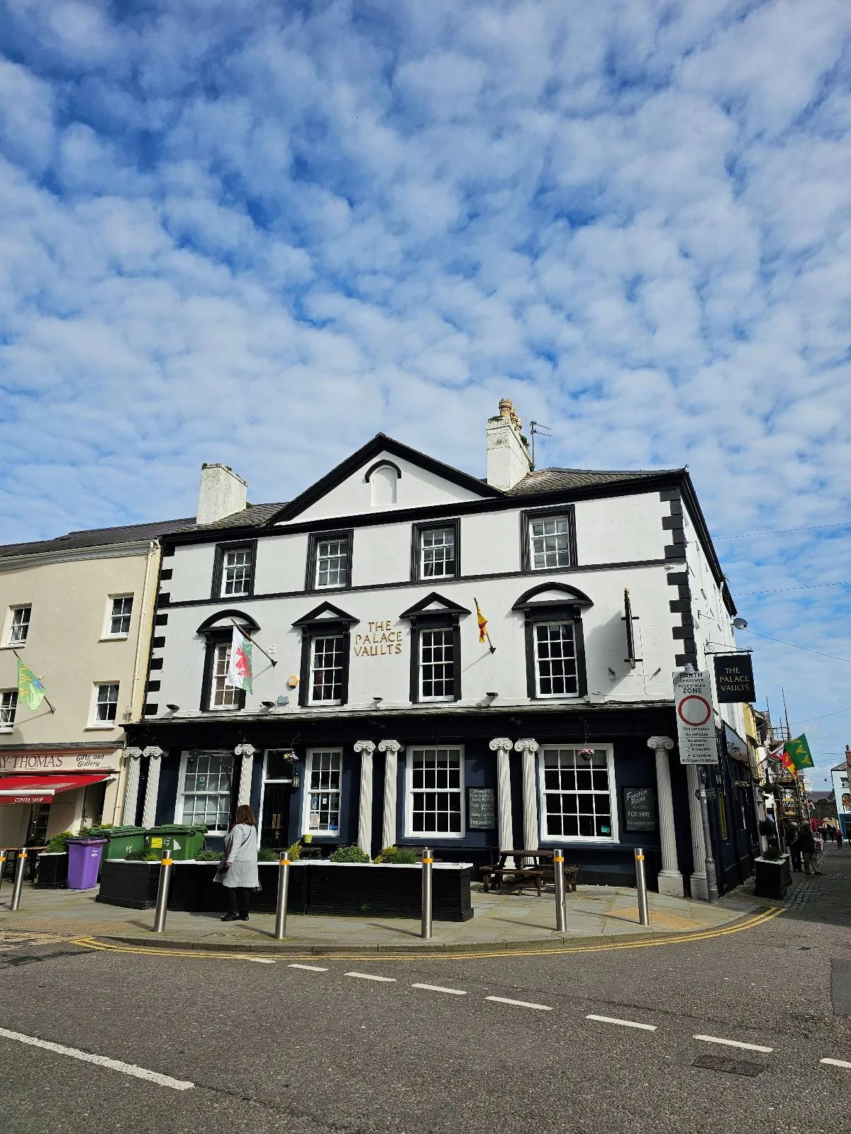 A white and black three-story building with large front windows and columns stands on a street corner under a blue sky with scattered clouds. Nearby, other buildings and a few street signs are visible.