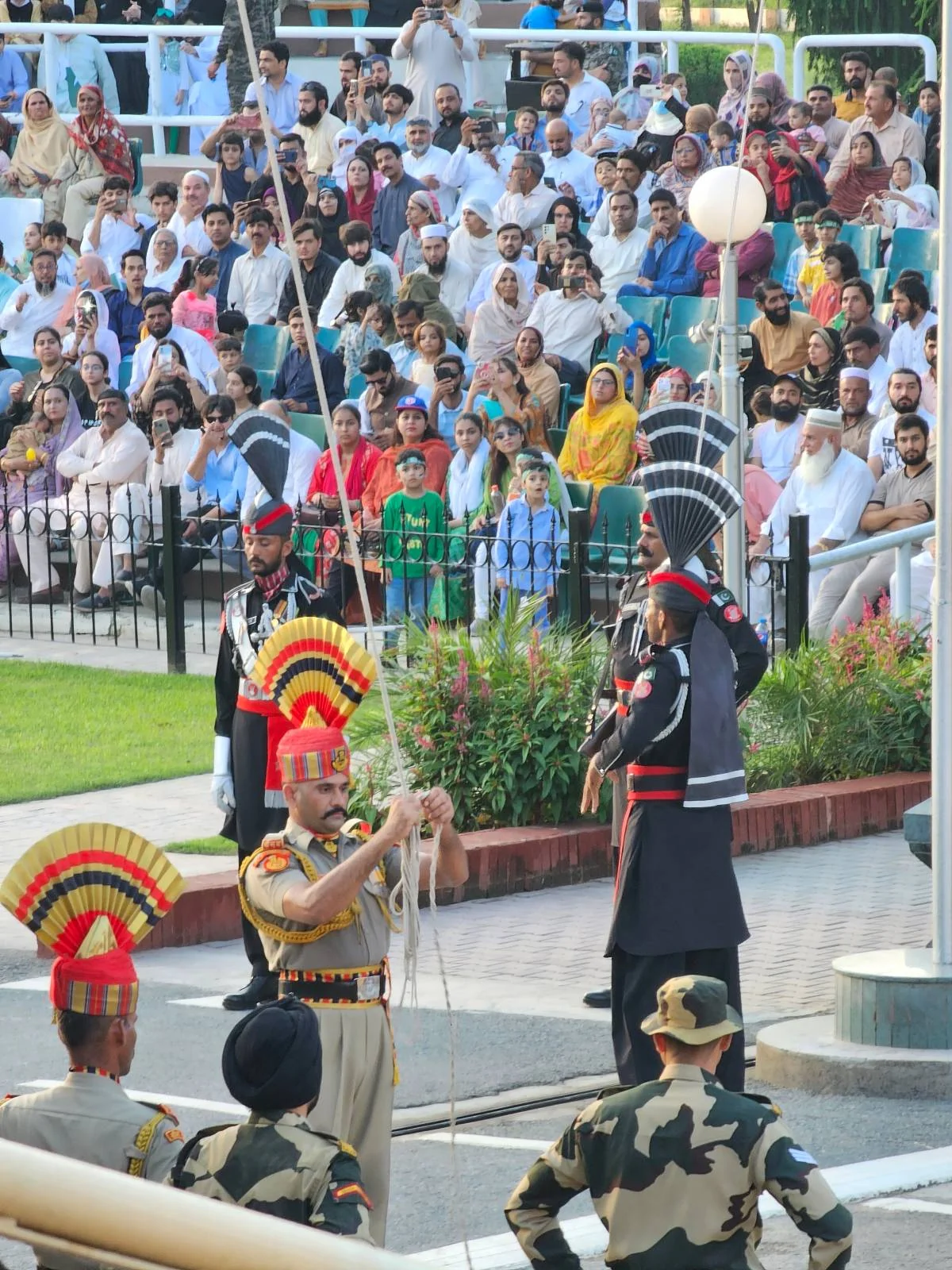Indian and Pakistani soldiers in ceremonial uniforms perform the Wagah border ceremony as a large crowd watches from the stands in the background. The scene is lively and colorful with flags and military regalia.