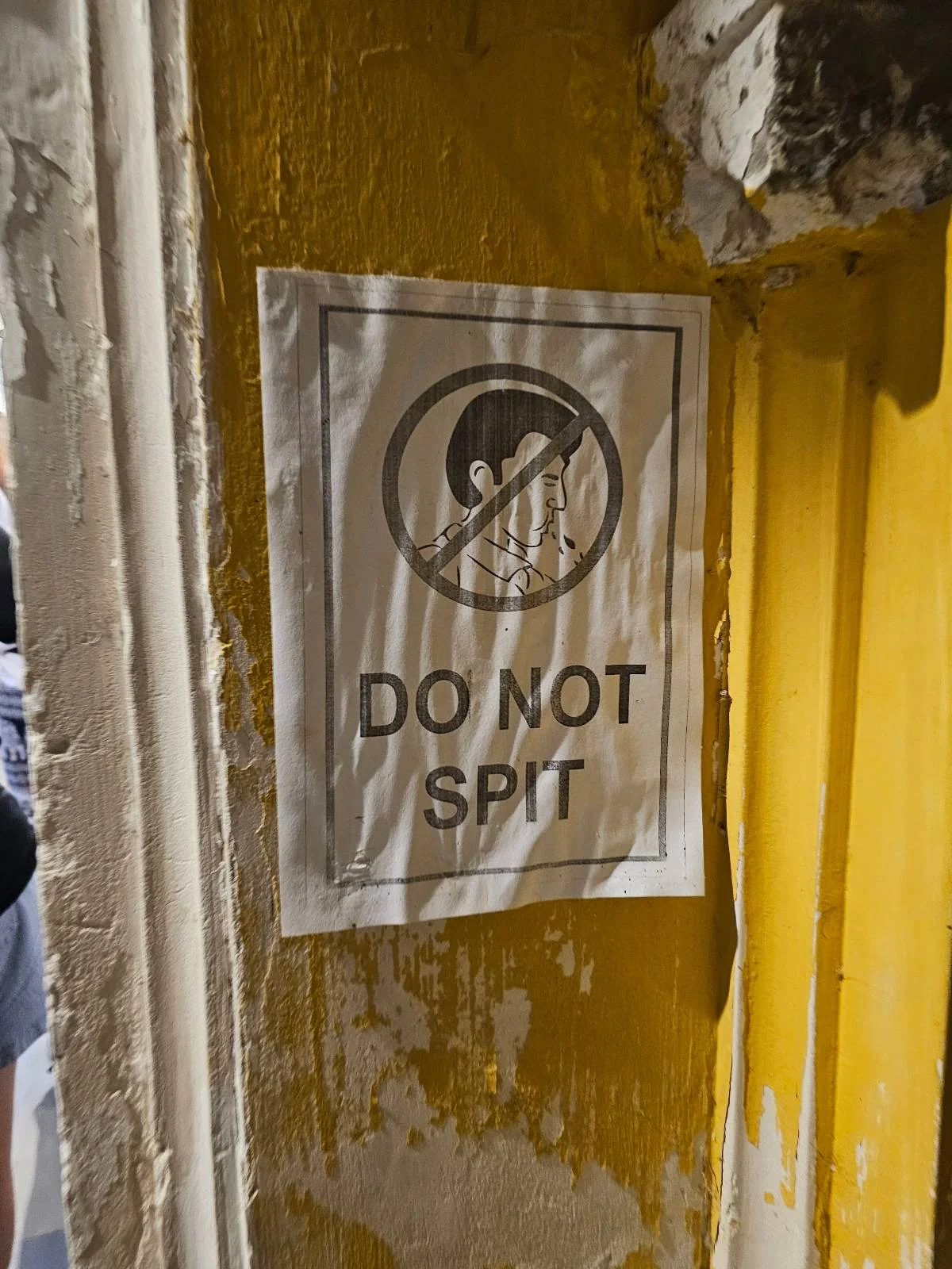 A white sign on a yellow, weathered wall displays a graphic of a person spitting with a prohibition symbol and the text "DO NOT SPIT" in bold letters.