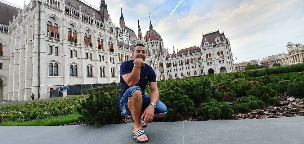 Man crouching and smiling in front of a historic Gothic-style building with green shrubs. Sky is partly cloudy, creating a cheerful mood.