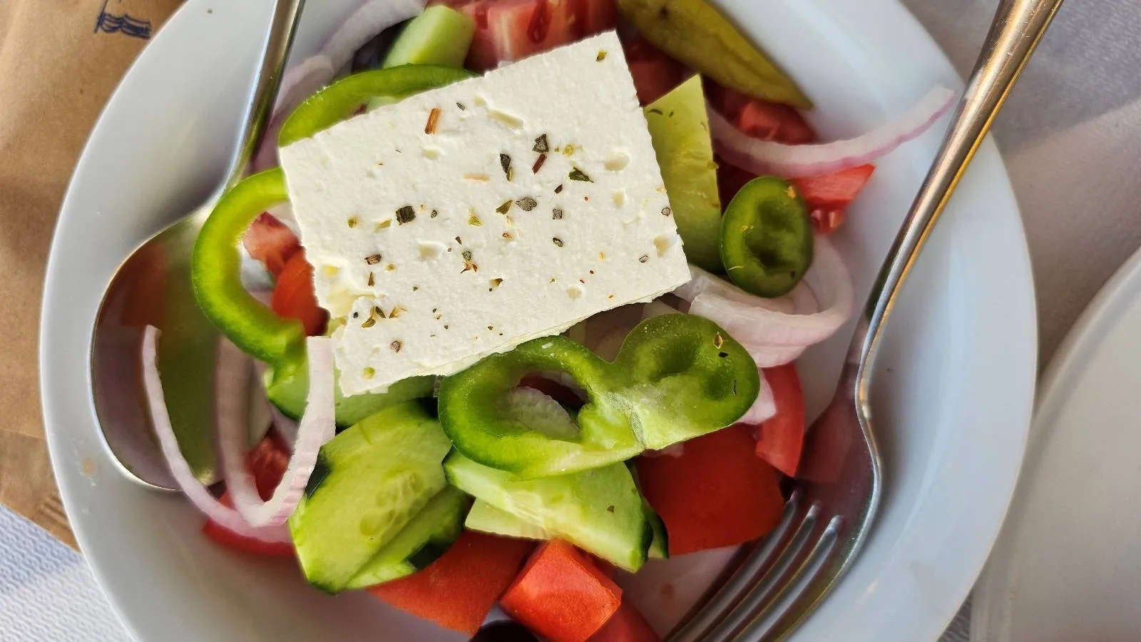A bowl of Greek salad with chopped cucumbers, tomatoes, green bell pepper, red onion, and a slice of feta cheese on top, garnished with herbs. A fork and spoon rest on the edge of the bowl.
