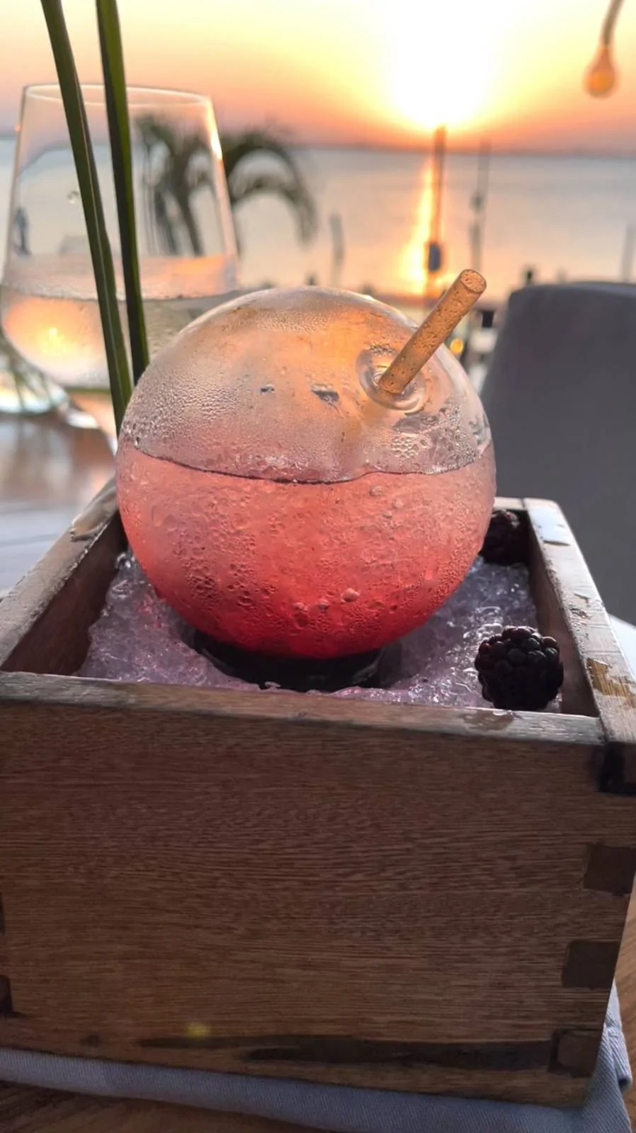 A spherical cocktail glass filled with a pink, icy drink sits in a wooden box with crushed ice. A straw is inserted into the drink. In the background, there's a sunset view over the ocean with blurred plants and wine glasses.
