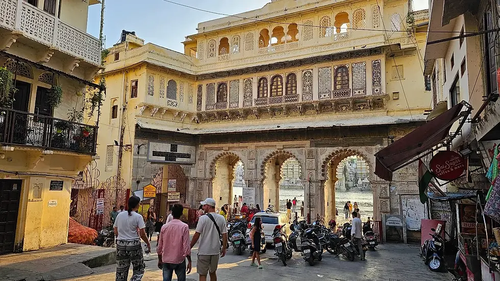 People walk towards a grand arched gateway in a historic building. Motorbikes line the street. Sign reads "Sai Sagar." Bright, warm atmosphere.