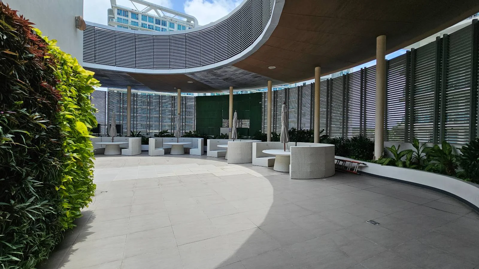 A modern outdoor seating area with circular benches and tables, surrounded by tall glass panels, green plants, and a partial roof letting sunlight in. A building is visible in the background under a blue sky with clouds.