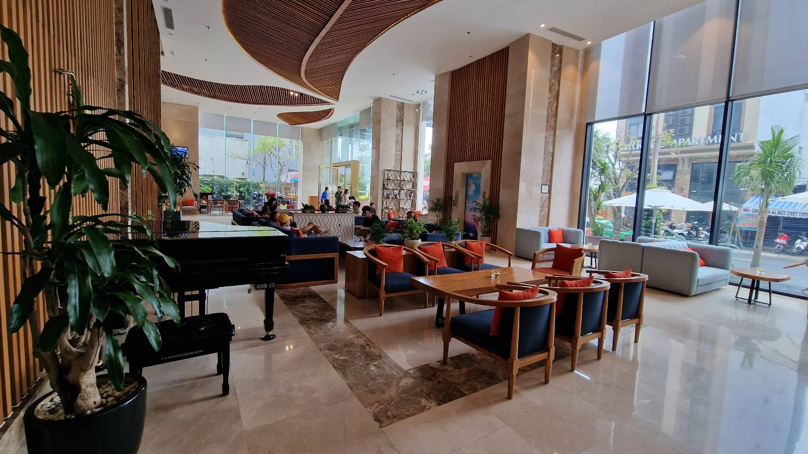 Spacious hotel lobby with large windows, a grand piano on the left, wooden furniture, and orange cushions. There are plants and artwork on the walls. Outside, trees and tables are visible through the windows.