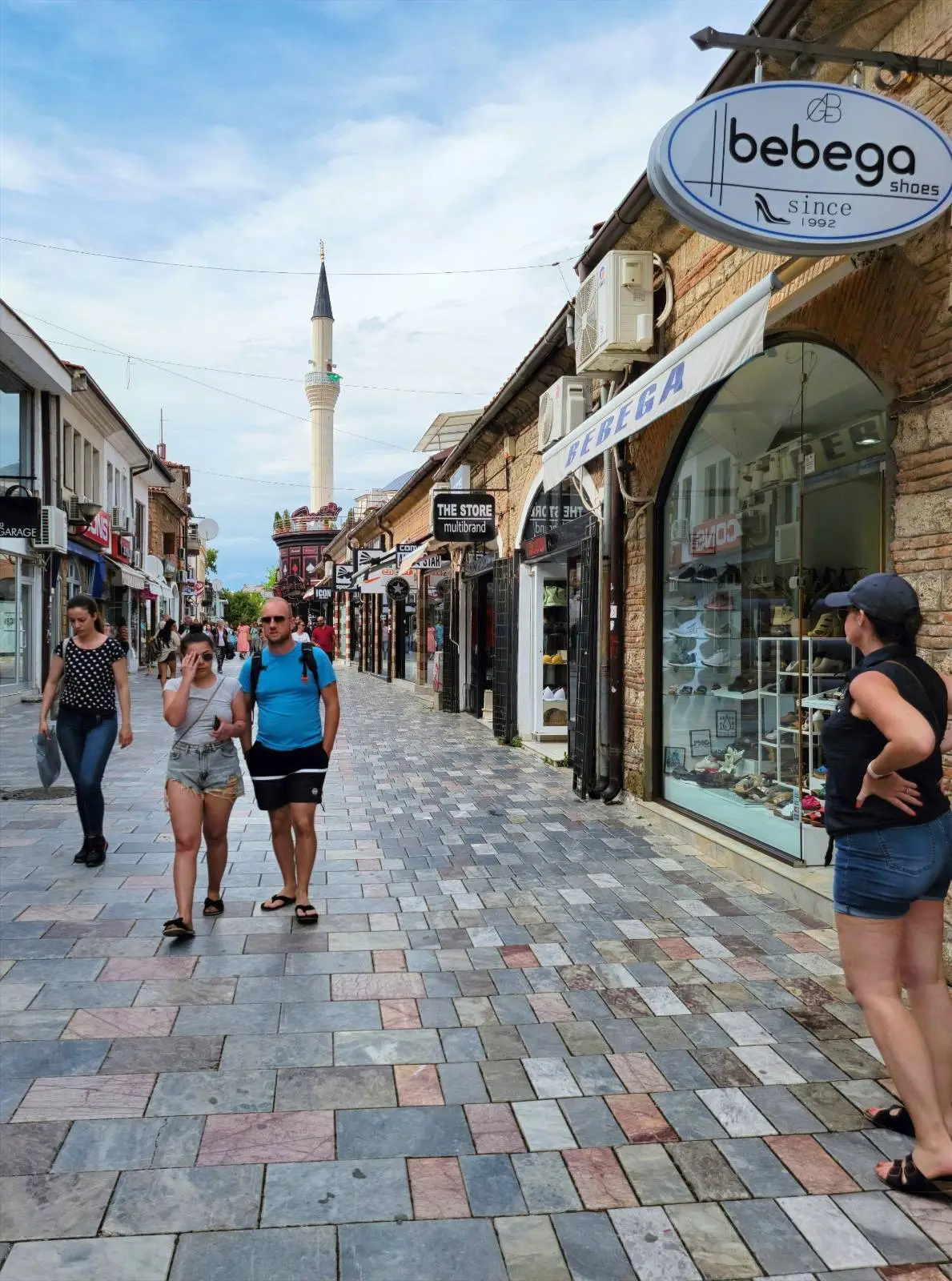 A narrow street with cobblestones lined with shops and a visible minaret in the background. Three people walk on the left, while a person stands on the right near a shop called "bebego." The sky is partly cloudy.