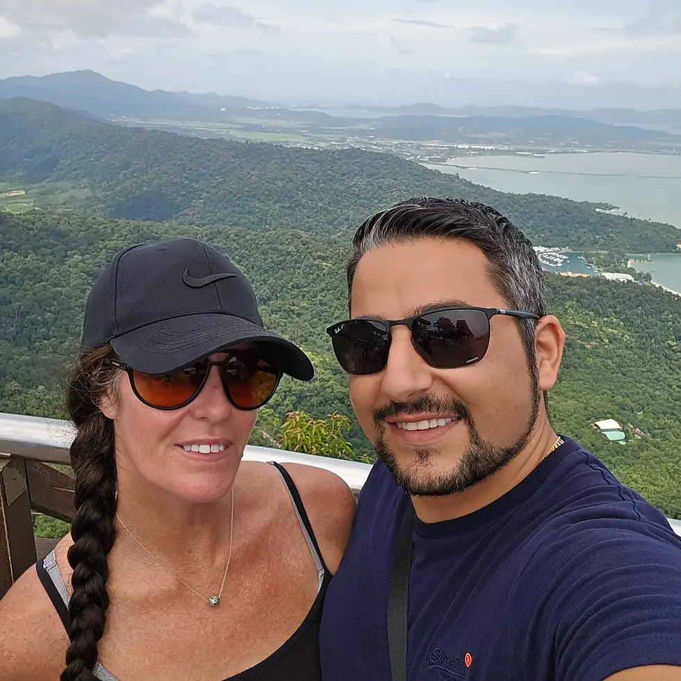 A man and woman smiling while taking a selfie against a lush, mountainous landscape. Both wear sunglasses; the sky is cloudy.