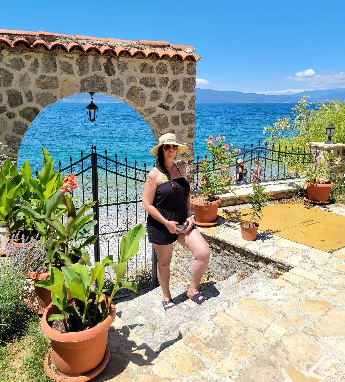 Person in a sun hat and black outfit stands by a stone archway overlooking a bright blue sea. The setting includes potted plants, a gated entrance, and a sunny sky.