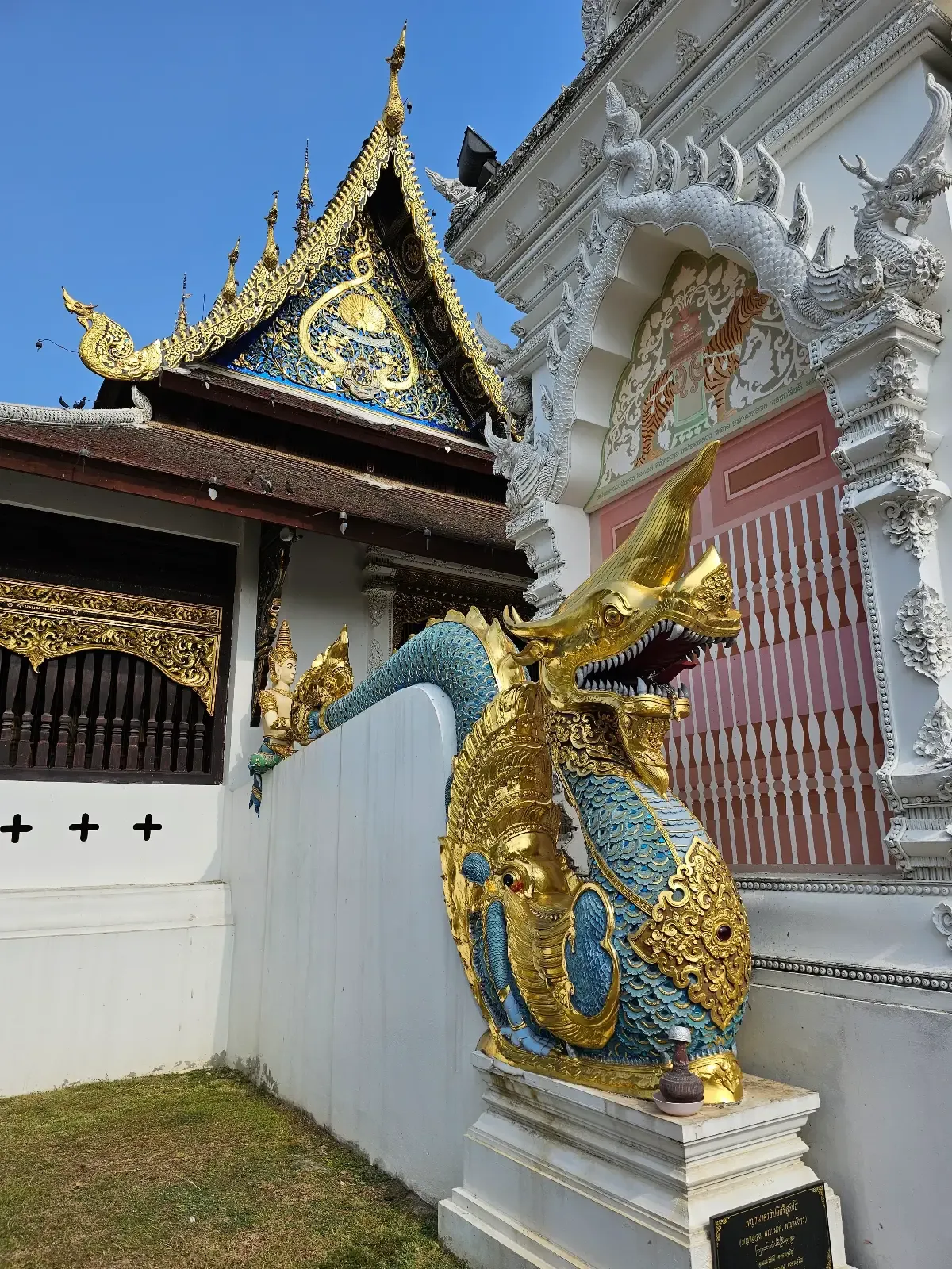 A traditional Thai temple features an ornate gold and blue dragon sculpture along a white wall, with intricate roof and architectural details visible in the background.