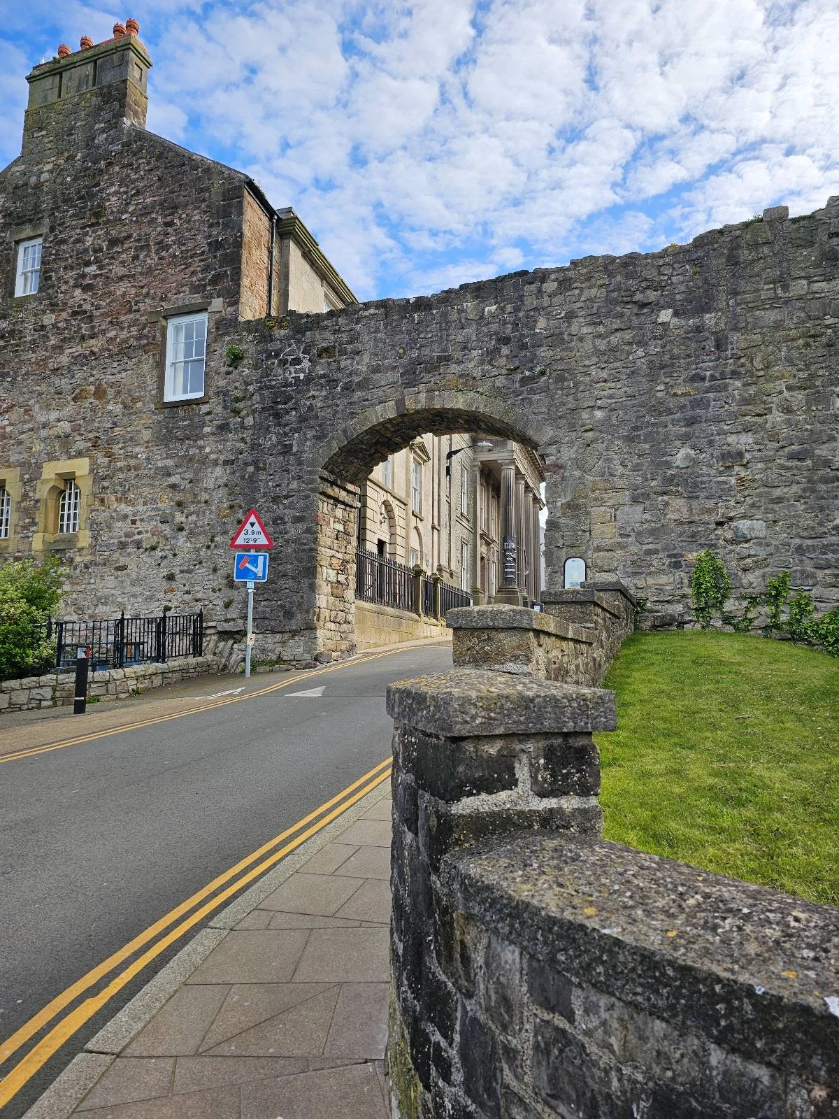 A stone archway built into an old stone wall stands over a narrow road with yellow lines. The sky is blue with scattered clouds, and green grass grows beside the wall.