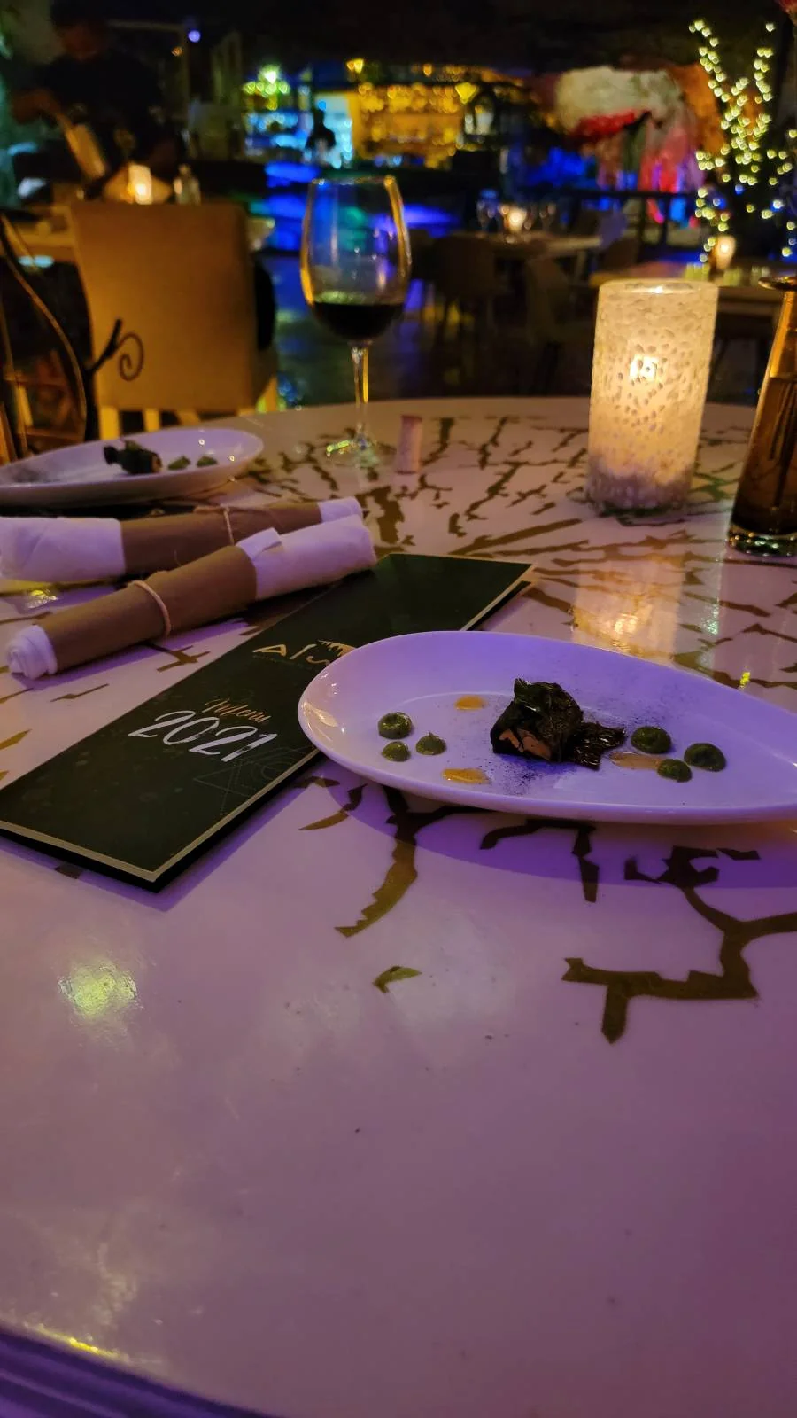 A dimly lit restaurant table with a white plate containing a small dish, accompanied by a menu. A candle in a glass holder and a wine glass are also on the table. The background features colorful lights and blurred decor.