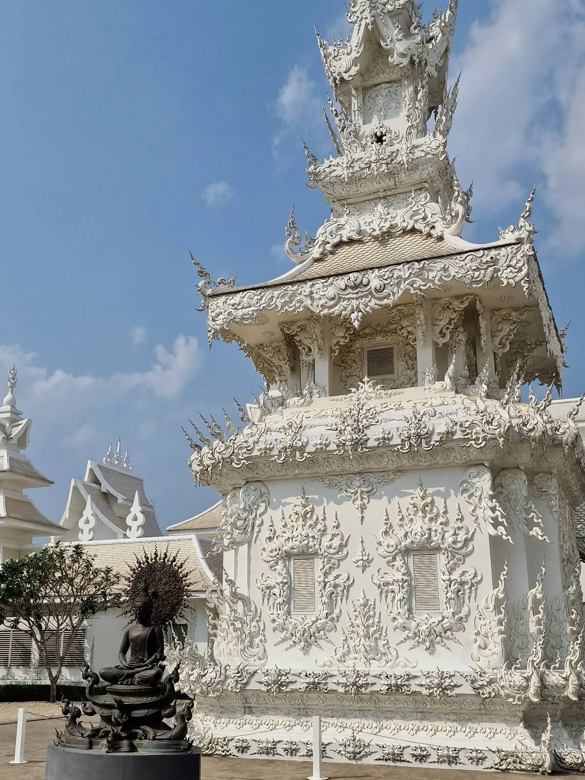 A beautifully ornate white temple with intricate carvings and details, set against a clear blue sky. In the foreground, there is a dark statue, adding contrast to the bright structure. Lush greenery is visible in the background.