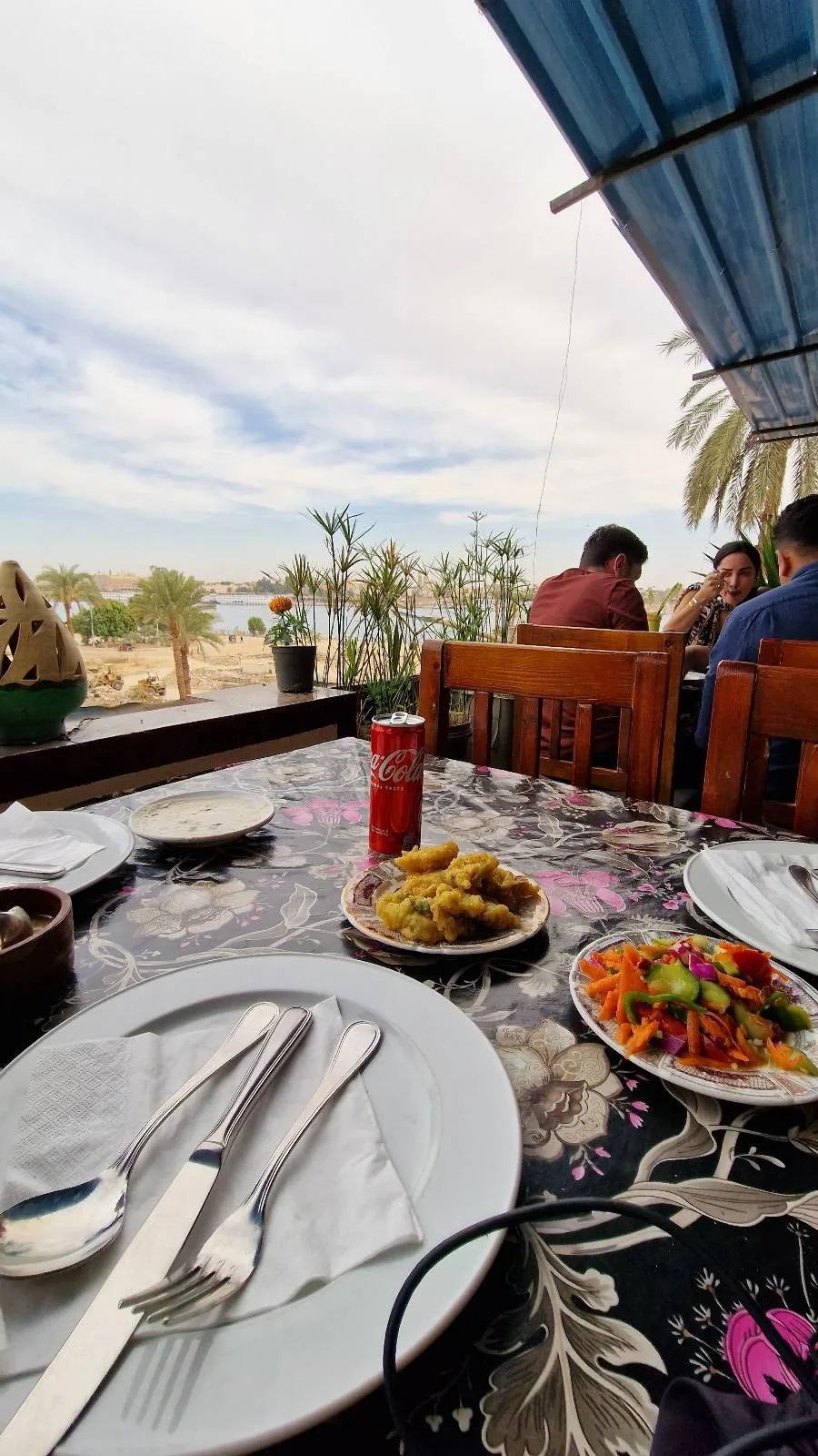 Outdoor dining setup with plates, cutlery, a small dish of mixed vegetables, and a basket of bite-sized snacks on a decorated table. People are seated in the background under a canopy, with palm trees and a bright sky visible.