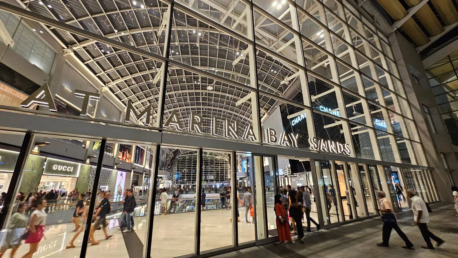 A large glass entrance to a shopping center with the words "Marina Bay Sands" displayed above. People are walking in and out, and the interior is brightly lit, showcasing various store signs.