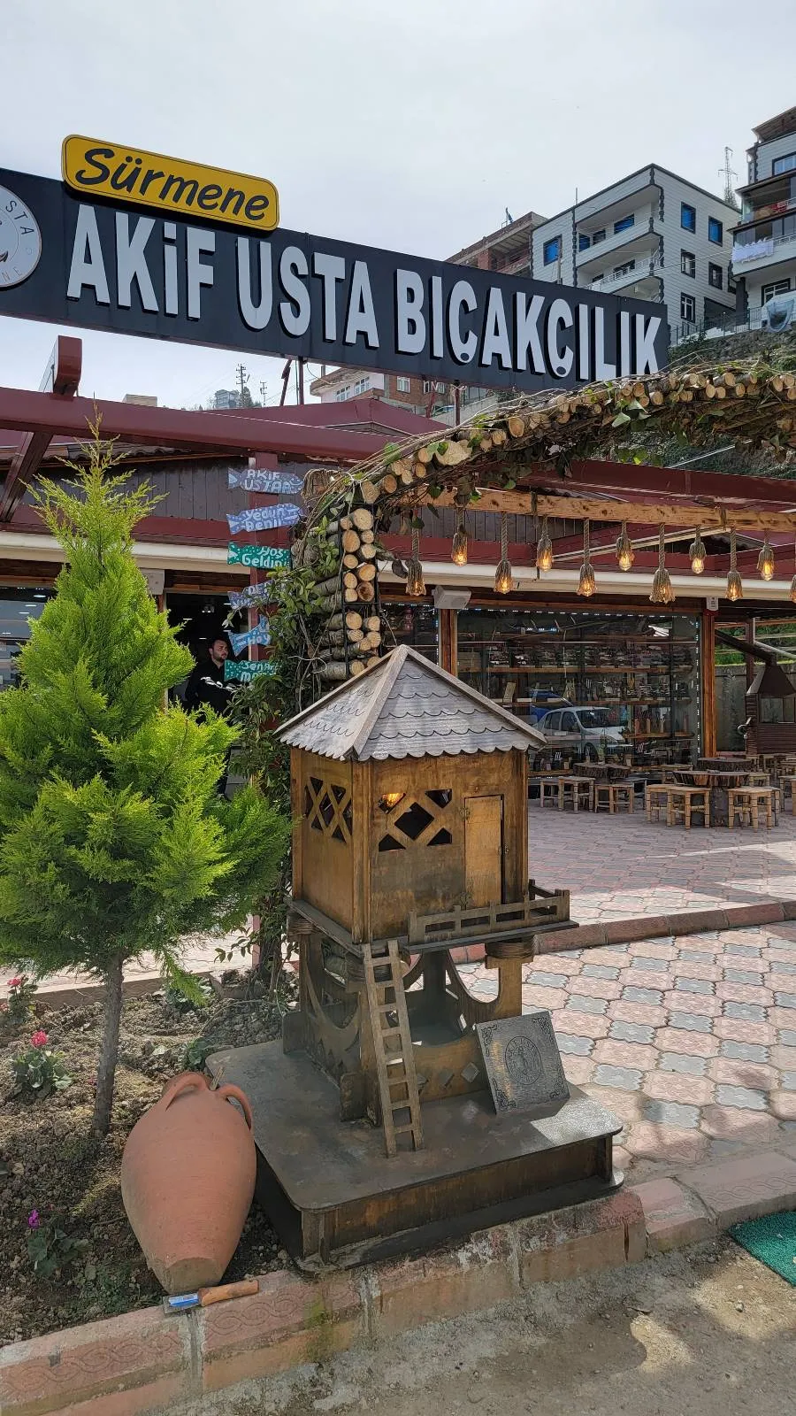 Outdoor area with a wooden structure and a sign overhead reading "Sürmene Akif Usta Bıçakçılık." A small tree and a large clay pot are visible in the foreground.
