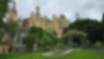 The historic Schwerin Castle, with a mix of architectural styles stands amid lush green gardens, with a cloudy sky overhead. Trees and manicured lawns surround the structure.
