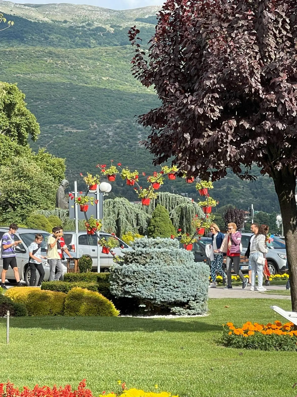 People walking in a park with vibrant flower arrangements and lush greenery. A large tree shades part of the scene, and mountains are visible in the background. Brightly colored flowers and shrubs decorate the park.