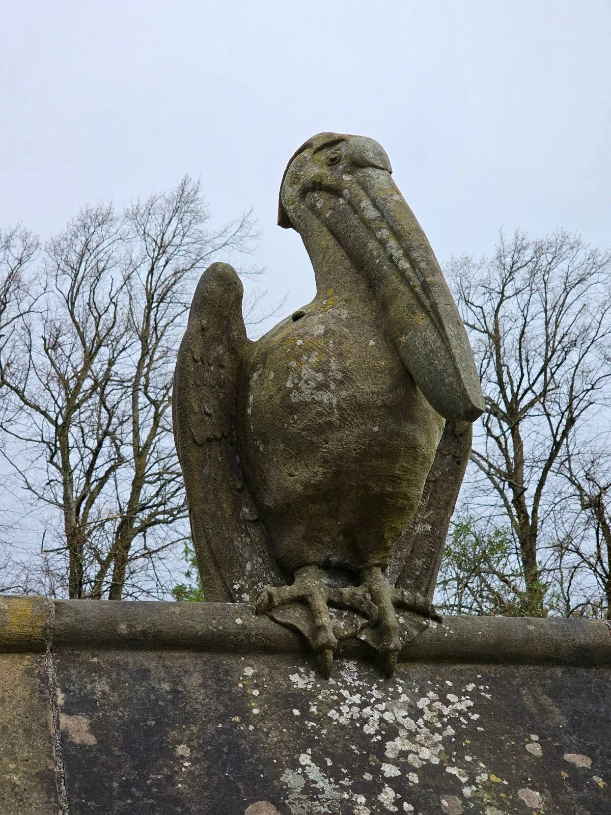 Stone sculpture of a pelican perched on metal railing, with leafless trees in the background.