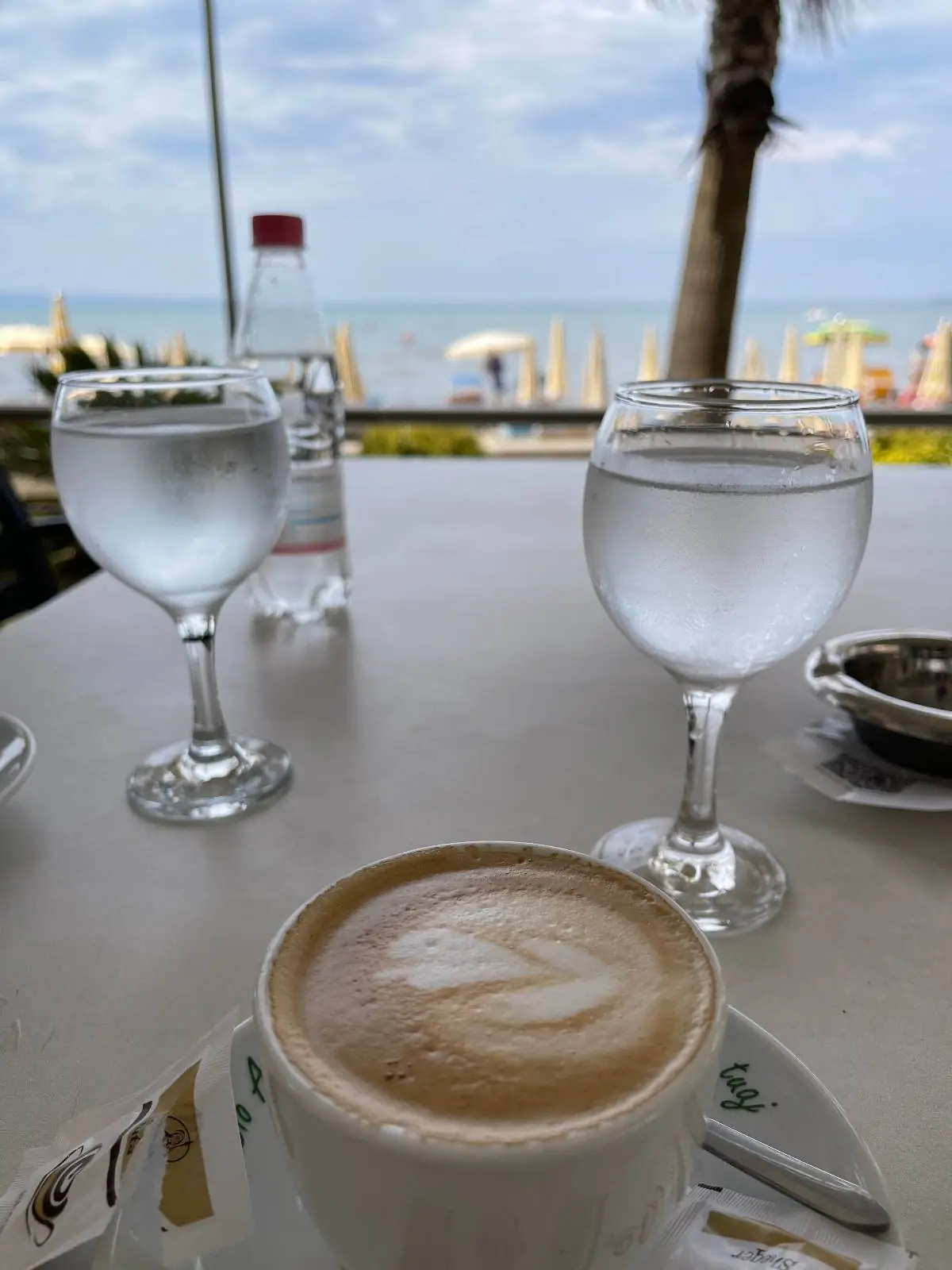 A cup of cappuccino and two glasses of water sit on a table overlooking a beach, with the ocean and umbrellas visible in the background under a cloudy sky.