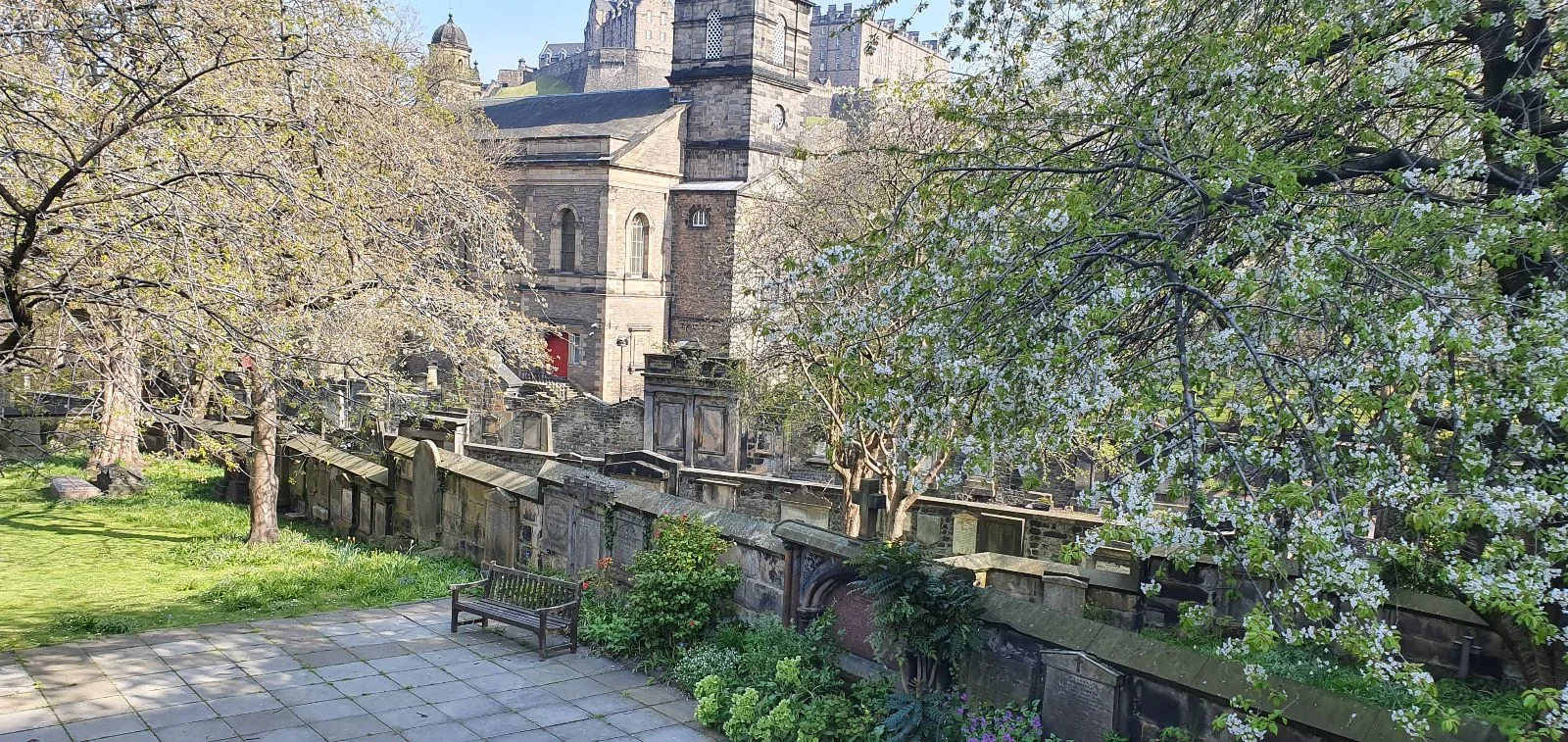 View of St Cuthbert’s Church and surrounding gardens in Edinburgh, with trees, stone walls, and a quiet seating area.