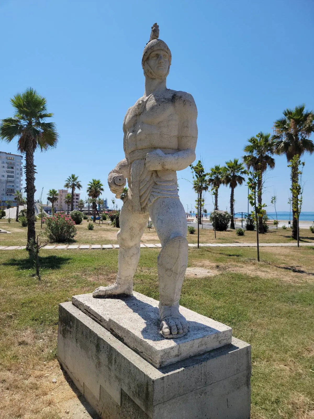 A stone statue of a warrior with a helmet stands on a pedestal in a park lined with palm trees, under a clear blue sky.