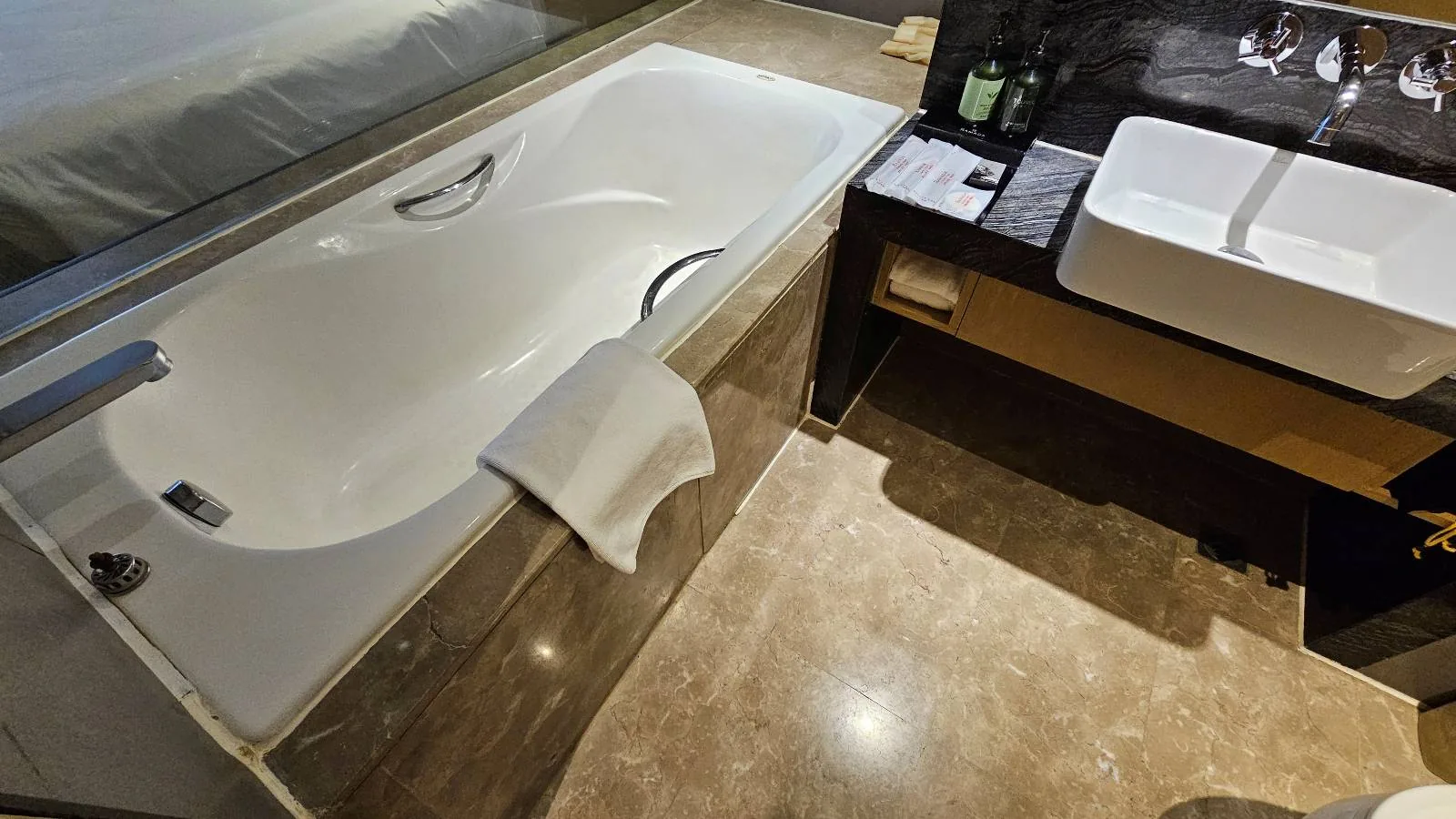 Modern bathroom with a white bathtub, a towel draped over the edge, a sink with a rectangular basin on a dark countertop, and toiletries neatly arranged beside it. Marble floor and glass partition are visible.