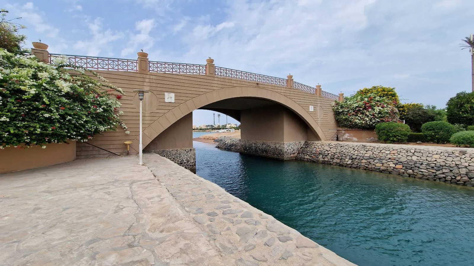 A stone bridge stretches over a man-made water channel, bordered by flowering shrubs and greenery on a partly cloudy day.