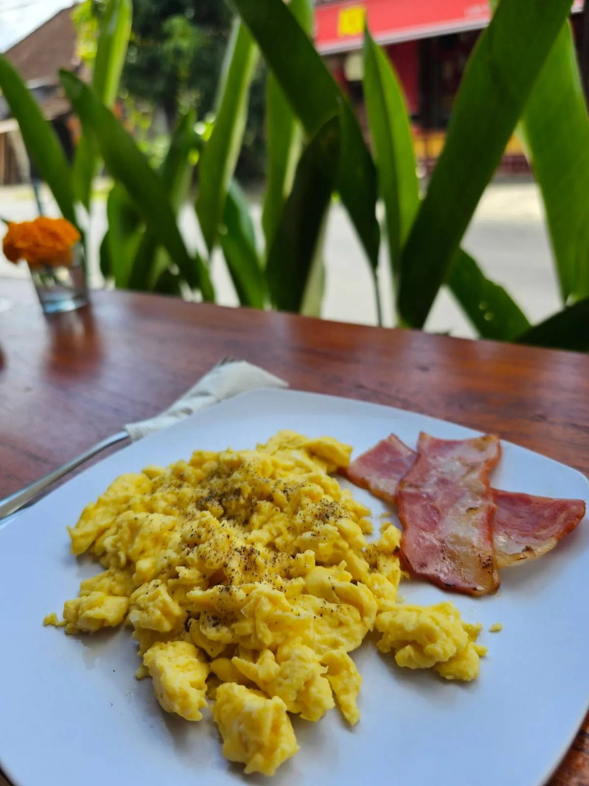 A white plate on a wooden table with scrambled eggs and two slices of bacon. In the background, green plants and an out-of-focus street view.