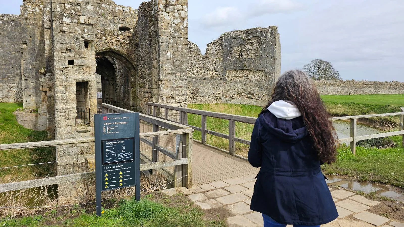 A person with long dark hair and a navy coat stands near a sign, facing the entrance of a stone castle ruin surrounded by grass and a wooden bridge.