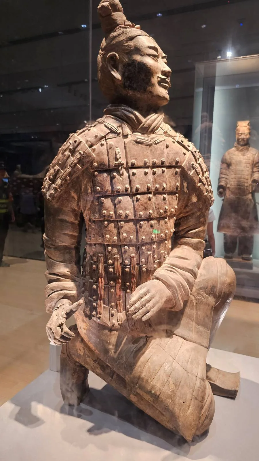 A terracotta warrior statue kneeling on one knee, dressed in detailed armor with a topknot hairstyle, displayed in a museum setting with other figures visible in the background.