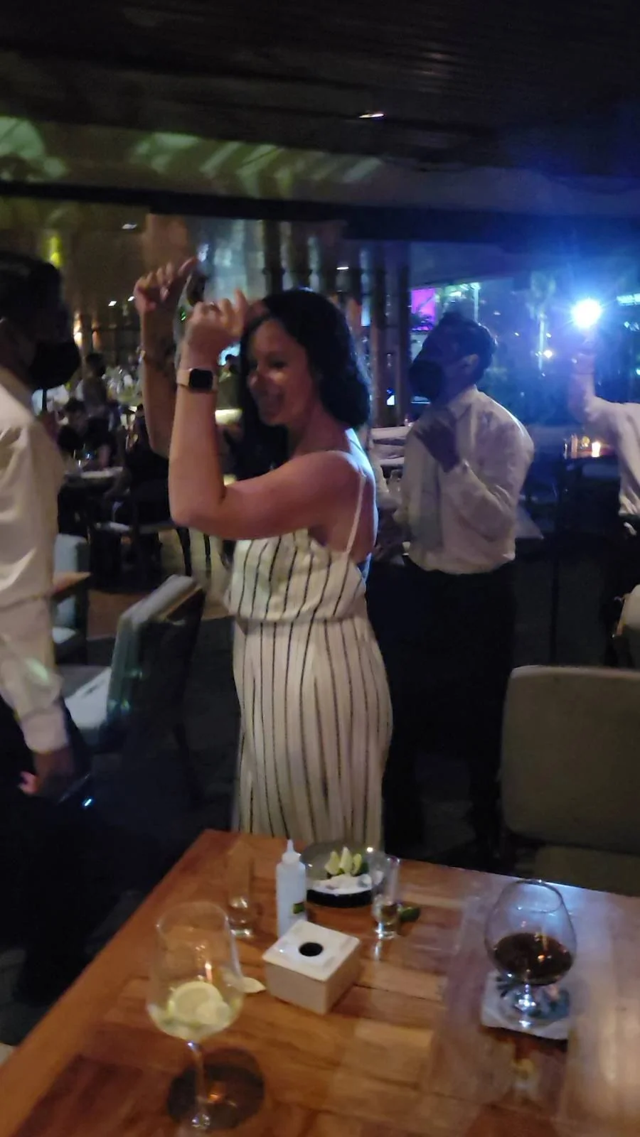 A woman in a striped dress is dancing in a dimly lit restaurant. Other diners are visible in the background. A table in the foreground holds a glass, a bowl of snacks, and other items. Colorful lights create a lively atmosphere.