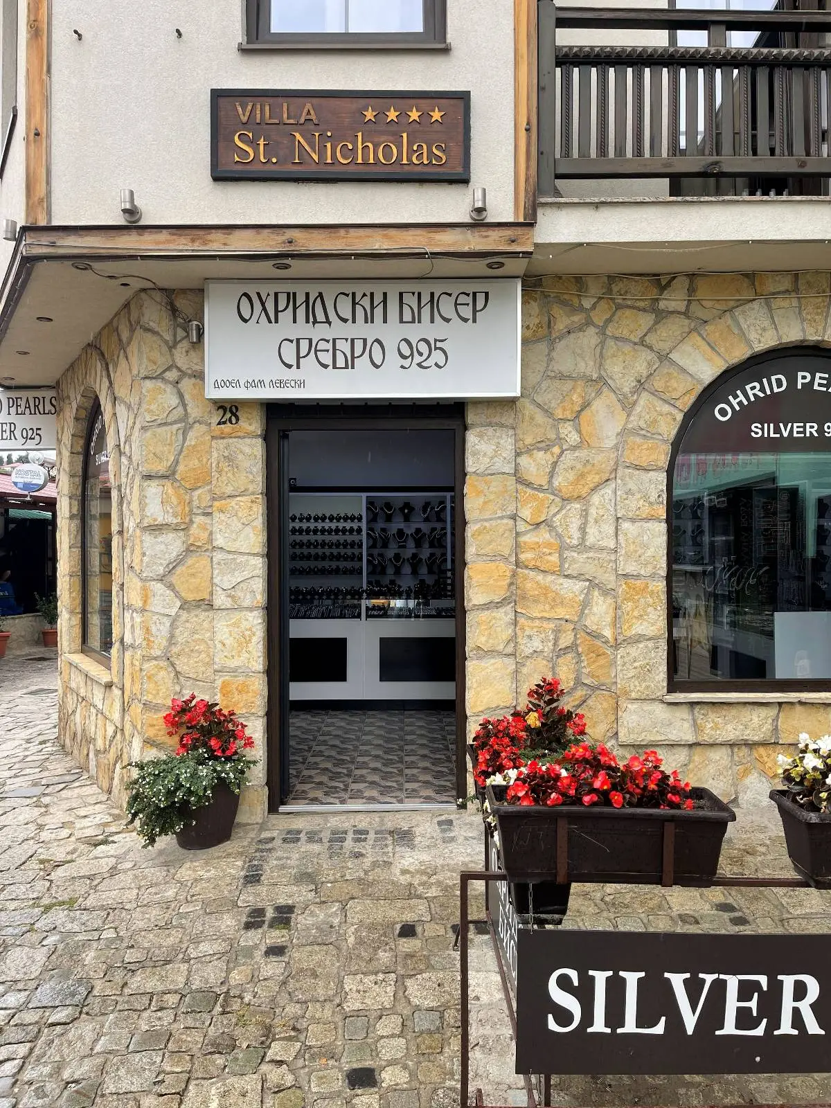 The image shows a stone building with a sign reading "Villa St. Nicholas." Below is another sign in Cyrillic. There is a doorway flanked by potted red flowers. The street is cobblestone, and a store window displays "SILVER" with jewelry inside.