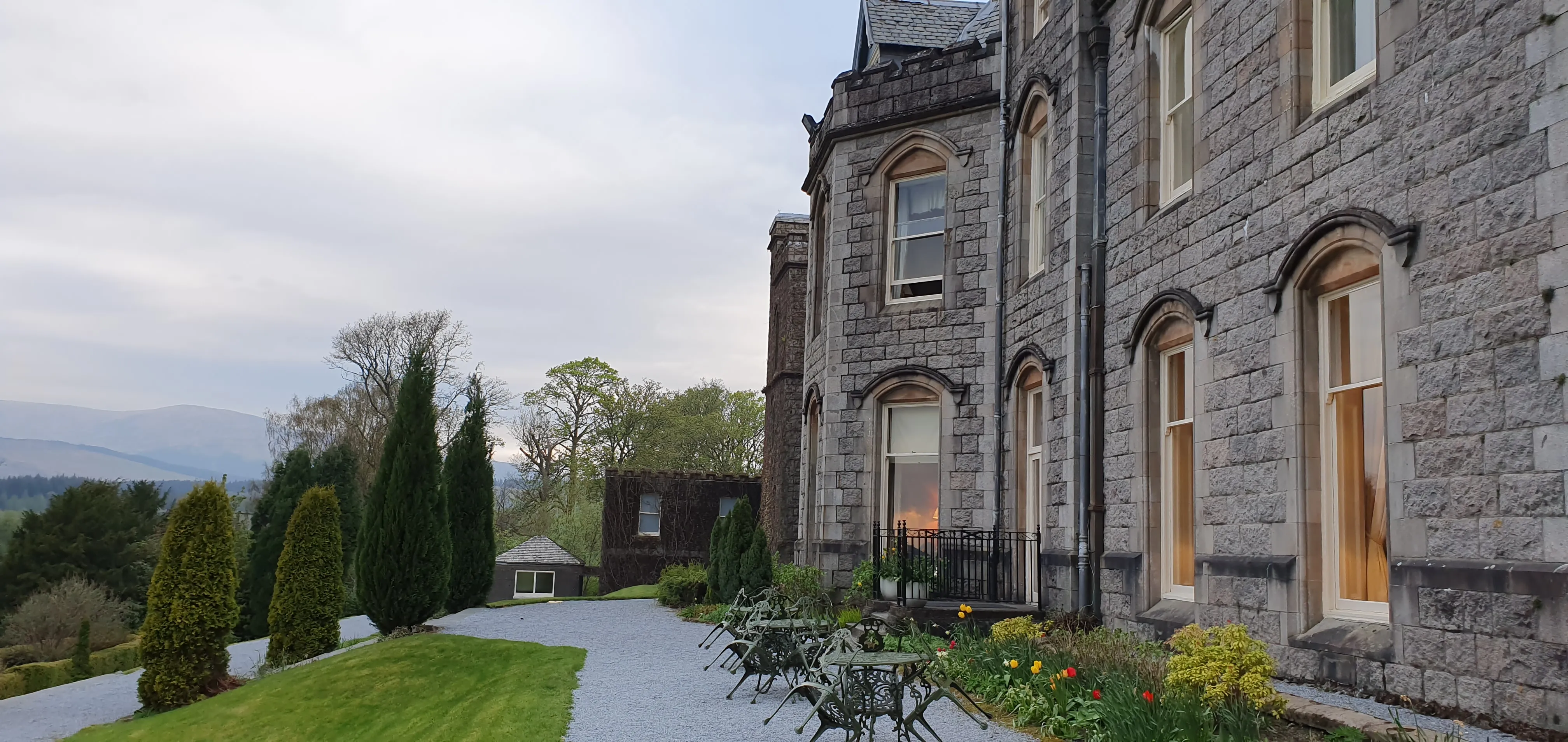 A grand stone castle-style luxury hotel with a clock tower, large windows, and a manicured green lawn under a cloudy Scottish sky.