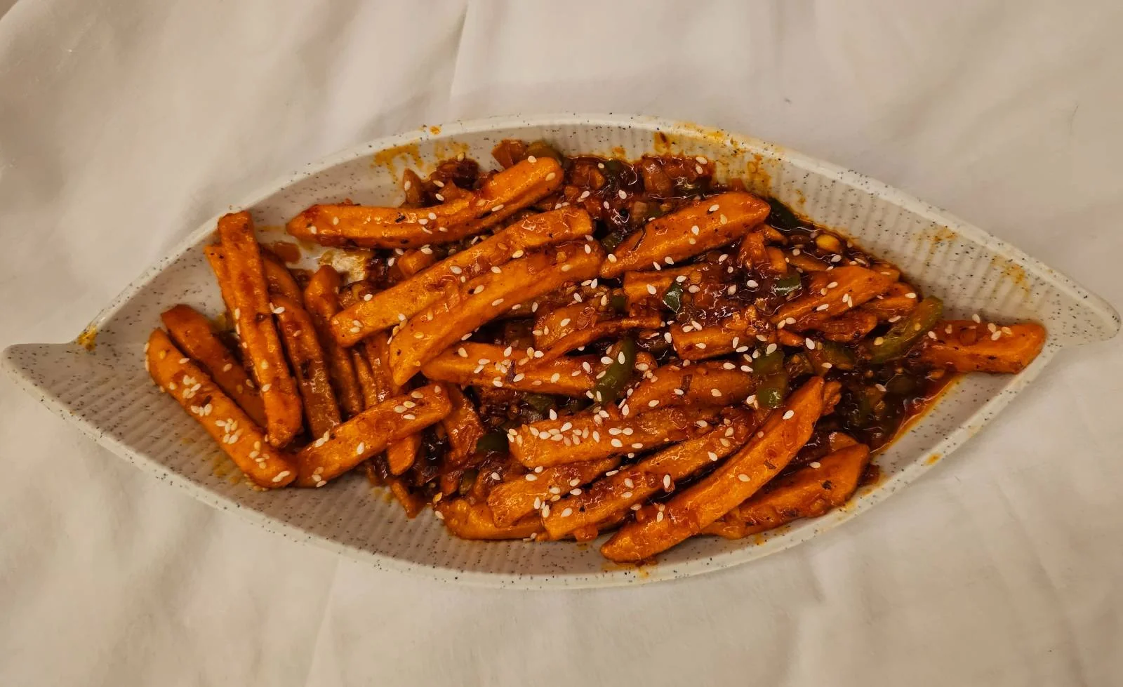 A plate of crispy orange sweet potato fries topped with seasoning and white sesame seeds, served on a white textured dish with a white fabric background.