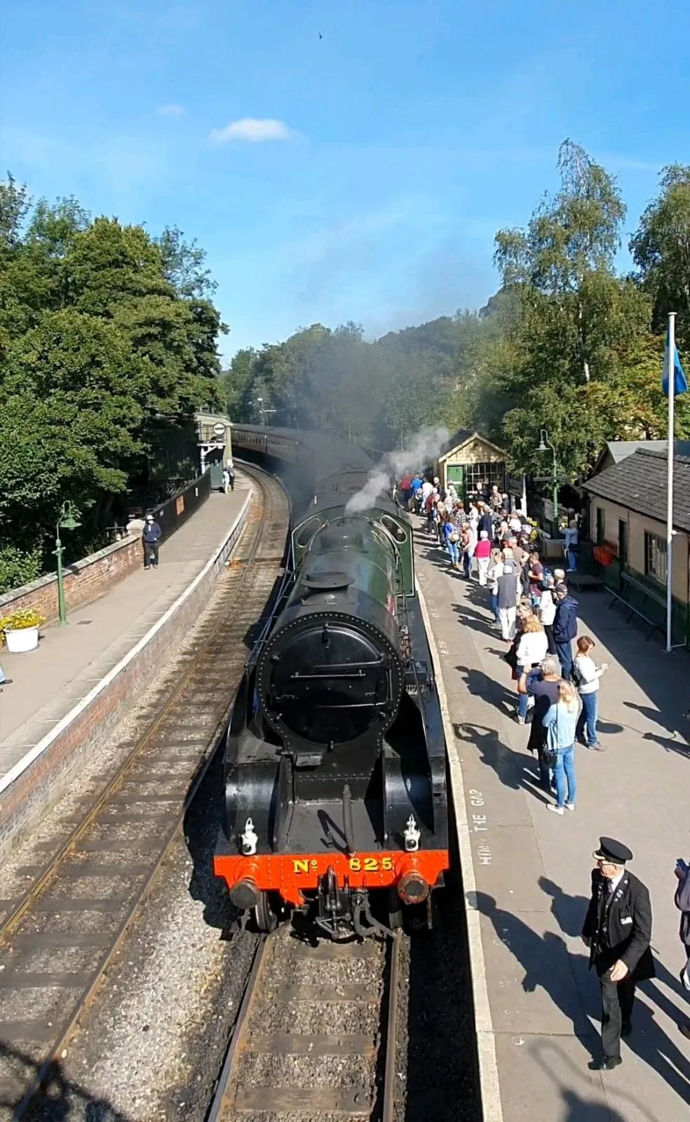 A black steam locomotive travels down a railway track under a blue sky. People stand on a nearby platform, watching the train go by. Trees line the background, and a few small buildings are visible.