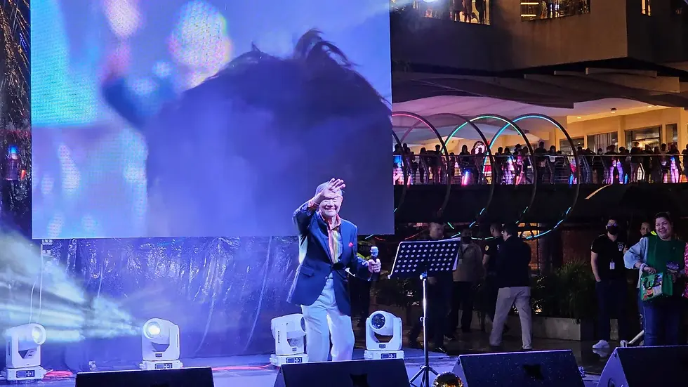 Man on stage waving, holding a microphone. Large screen projection behind. Crowd in the background. Bright stage lights and colorful accents.