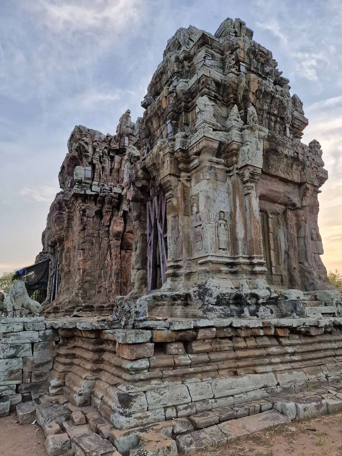 Ancient stone temple ruins with intricate carvings and weathered details, set against a partly cloudy sky at sunset. The structure appears partially eroded, showcasing historical architecture.